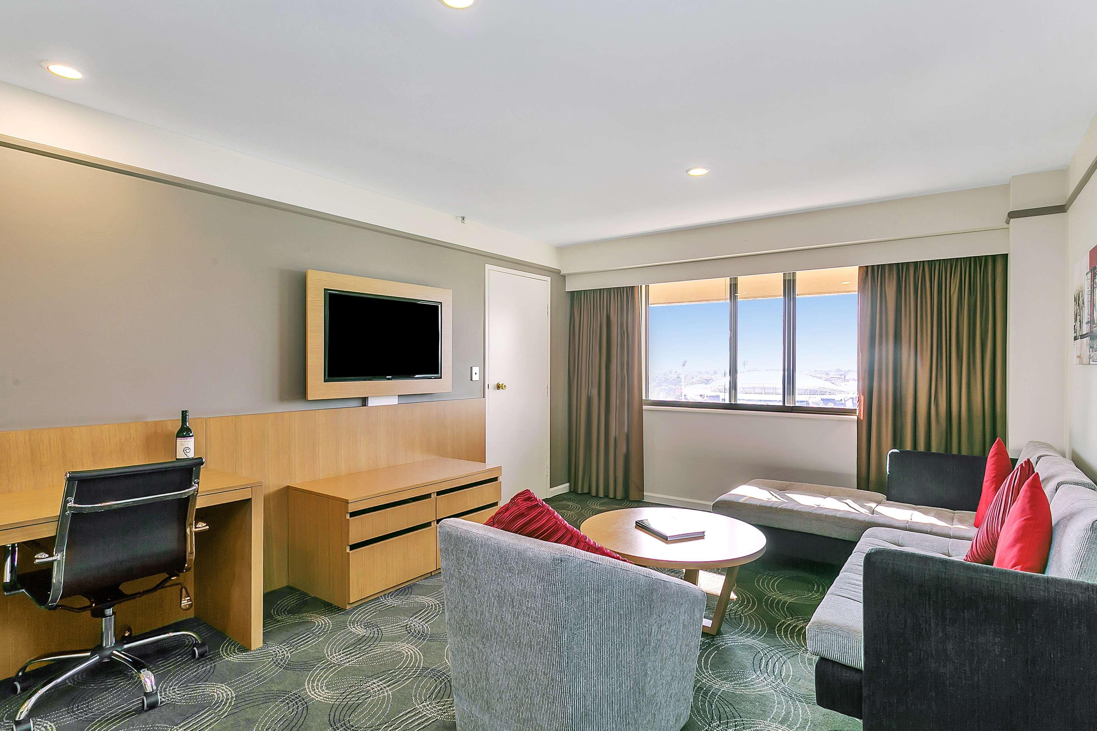 Stamford Plaza Adelaide Hotel Bagian luar foto The photo shows a modern hotel room or suite that features a cozy seating area. There is a comfortable sofa with red accent pillows, a round coffee table in the center, and a sleek desk with an office chair. On the wall, there is a flat-screen TV mou