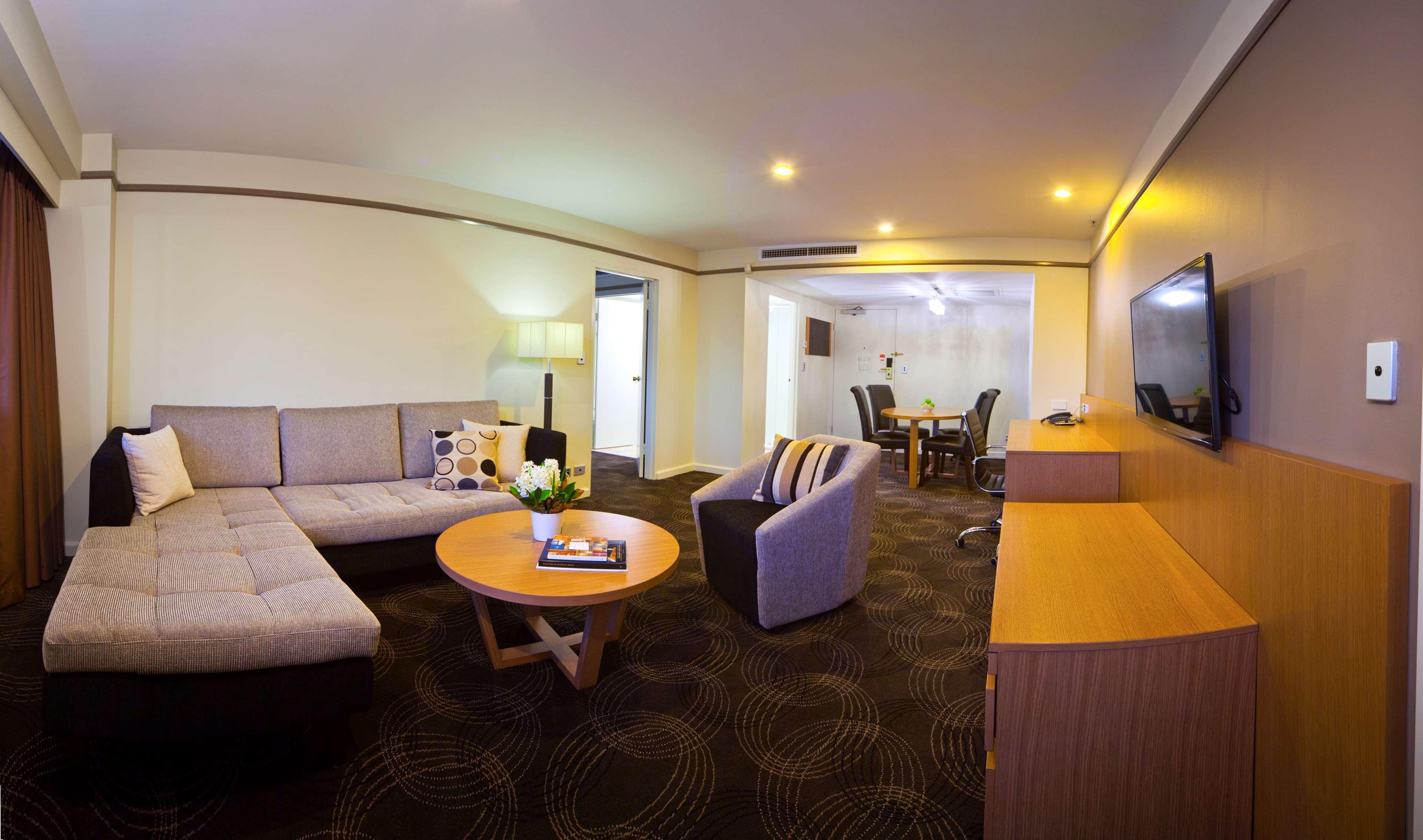 Stamford Plaza Adelaide Hotel Bagian luar foto The photo shows a spacious living area with modern decor. On the left, there is a light gray sectional sofa, accompanied by a round wooden coffee table in front of it. A couple of decorative pillows add a touch of color to the sofa. To the right, the