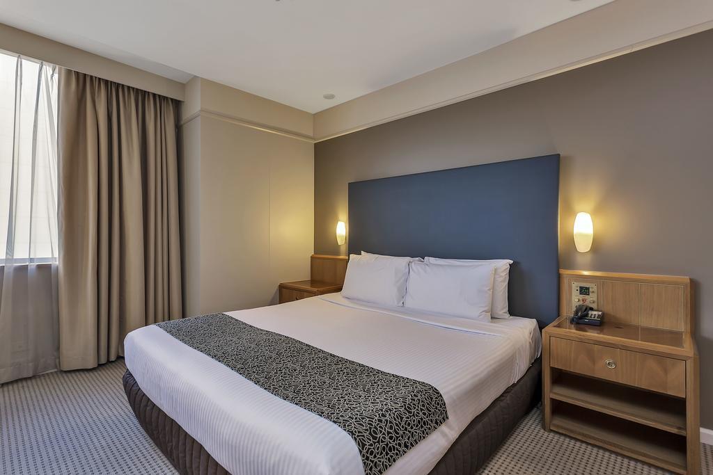 Stamford Plaza Adelaide Hotel Bagian luar foto The photo shows a modern hotel room. It features a double bed with a white duvet and a decorative grey and black throw. The headboard is a solid dark color. There are two bedside tables on either side of the bed, each with a lamp. Natural light enter